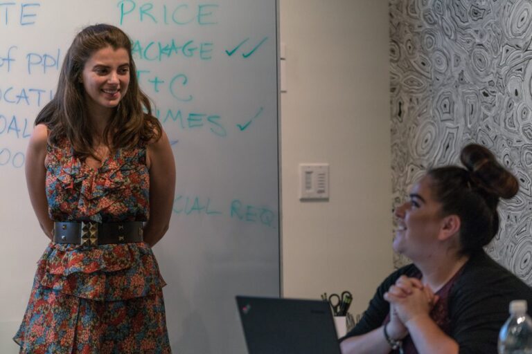 woman standing and smiling at another woman seated in front of a laptop