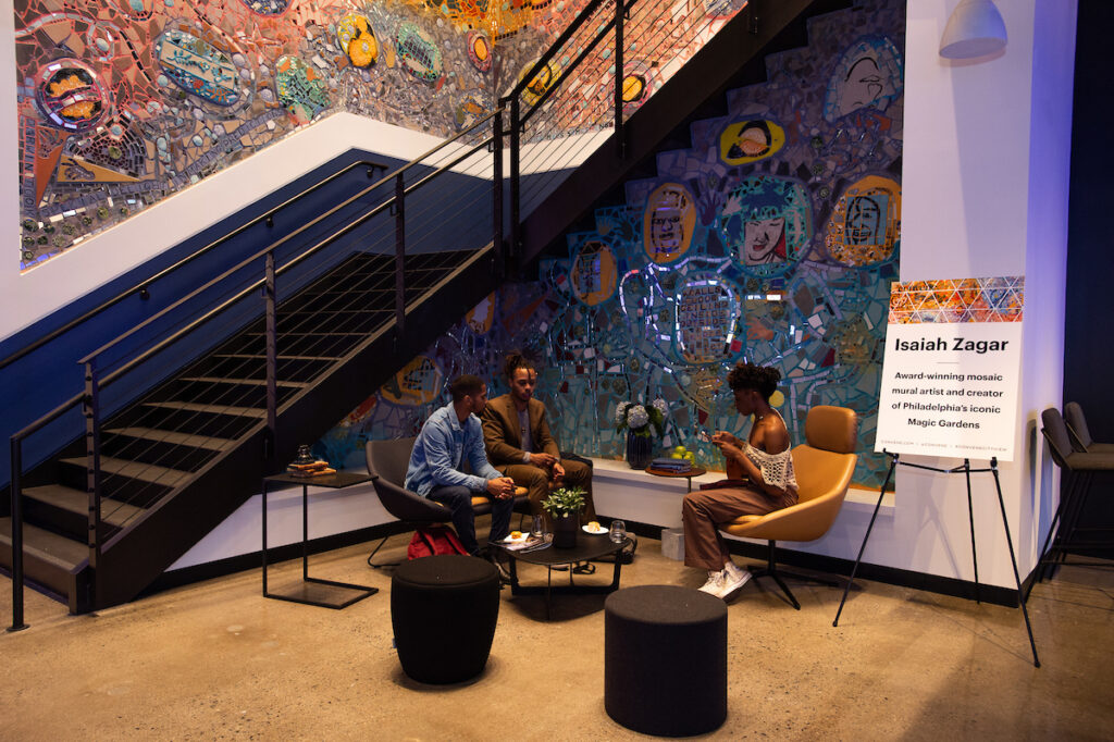 Three people sitting at the base of the stairs at Convene 30 S. 17th St. in contemporary furniture surrounded by glass and ceramic mosaic mural with sign saying "Isaiah Zagar."
