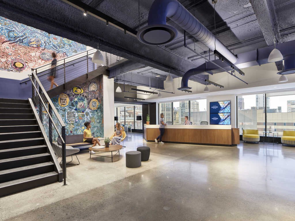 People in the welcome area of Convene S. 30 17th Street with large windows lighting a mixed media mosaic by Isaiah Zagar. 