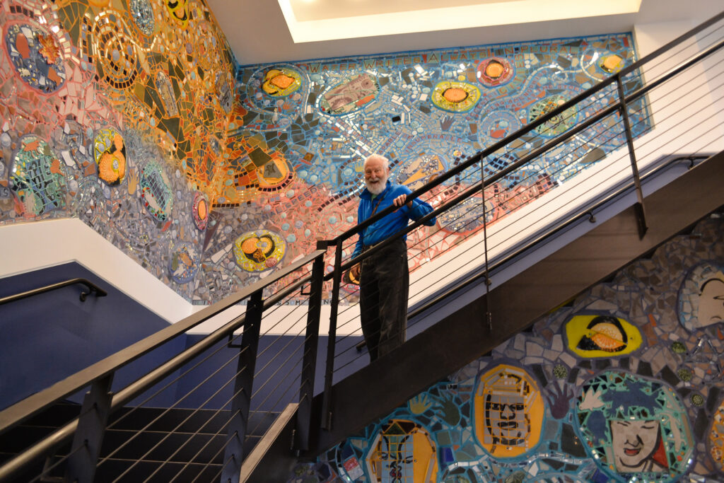 Artist Isaiah Zagar smiles while standing on the stair in front of a bright mosaic with faces and yellow circles visible in a large wavy pattern.