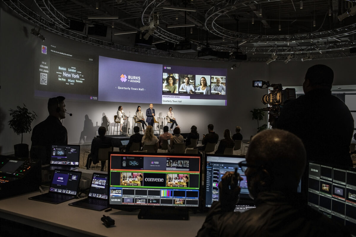 A large Convene meeting and event space. On stage there are a number of people who are presenting to a crowd.