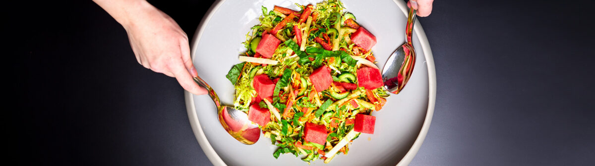 A Watermelon & Halloumi Salad in a large bowl. Someone is about to toss the salad with metal salad spoons.
