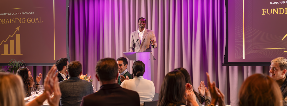 Presenter in front of audience with purple lighting and two screens.