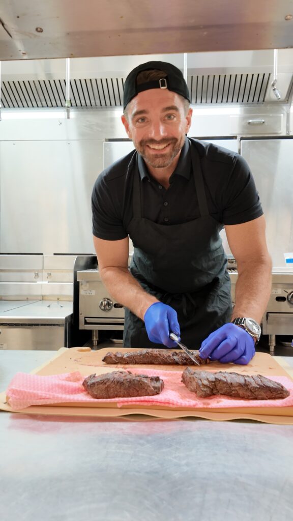 CEO Ryan Simonetti smiles as he slices steak in a commercial kitchen at Convene.