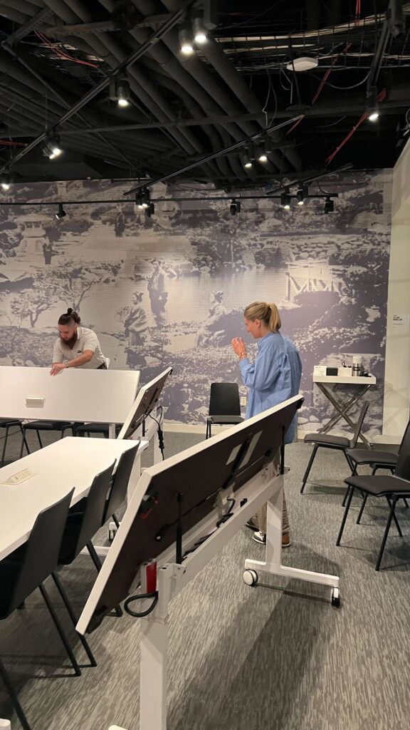 Two people setting up long classroom tables in Convene meeting room with mural.