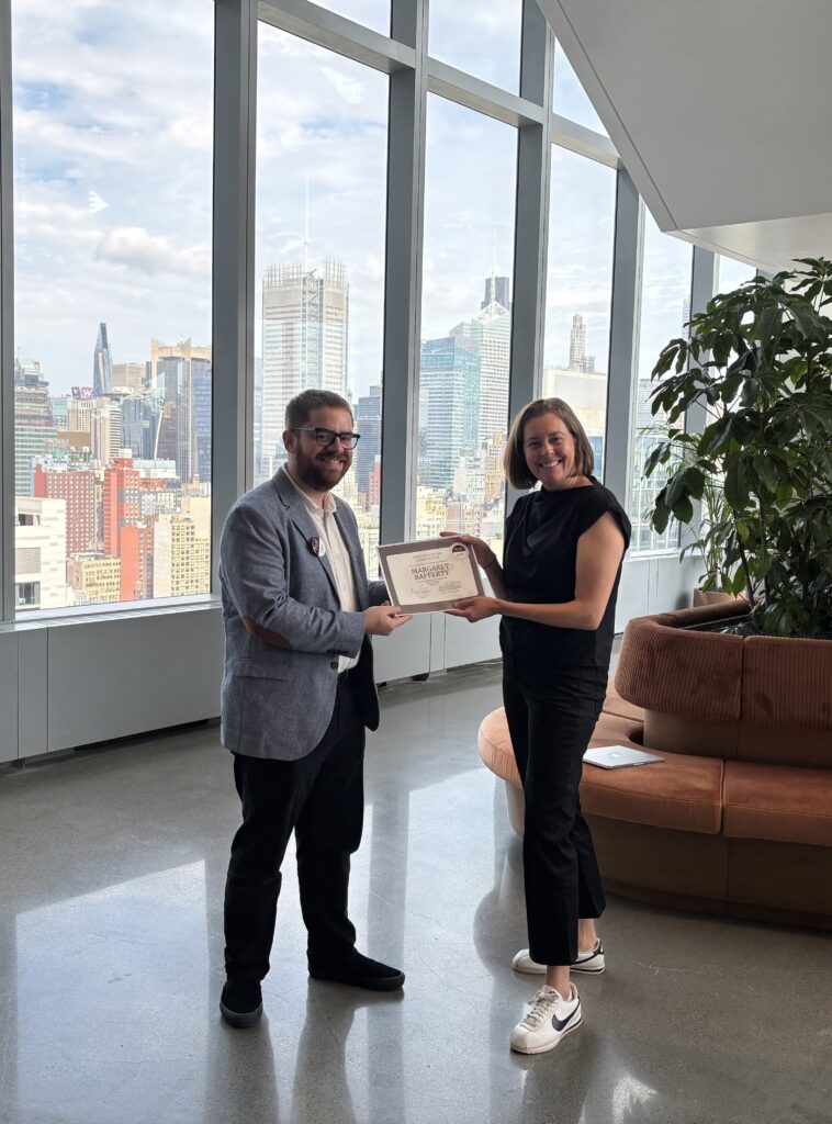 Person hands over a certificate to smiling person in front of NYC skyline.