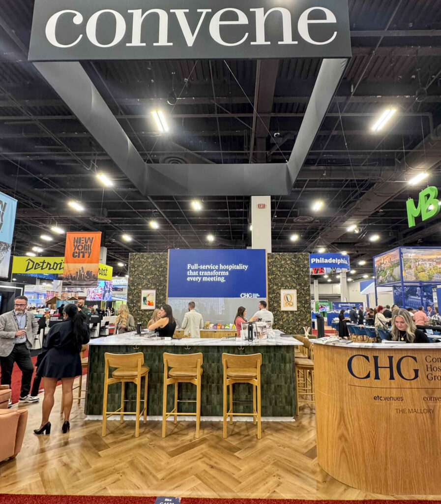 CHG booth on trade show floor at IMEX America with large sign saying "Convene" overhead. People are walking around a bar made with green tiles and multiple booths are visible.