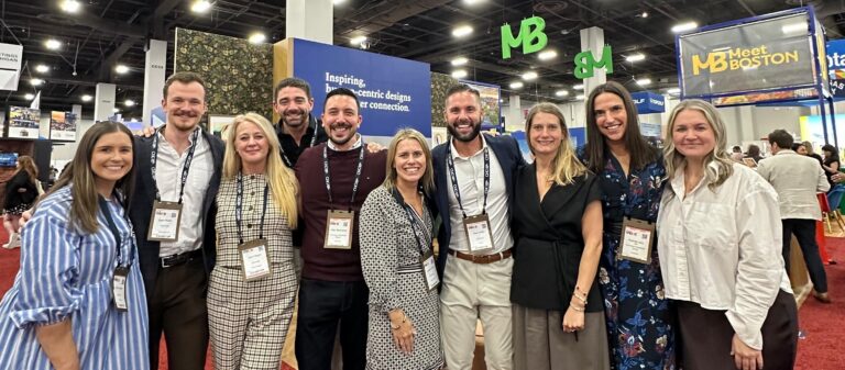 Team CHG at IMEX America smiling in line with booths in the background on a trade show floor.