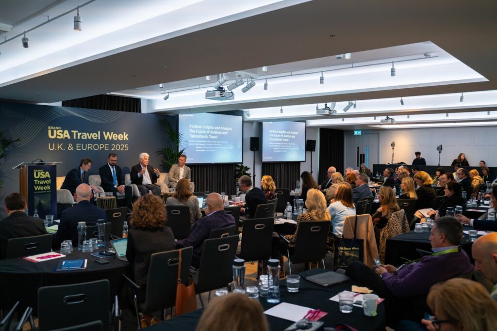 Group of panelist speaking to a seated crowd in a conference at etc.venues County Hall with banner for USA Travel Week on stage and projector screens.