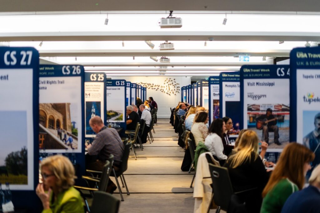 Rows of seated professionals inside the conference room at etc.venues County Hall in London. People are in separated booths for meetings with signage for U.S. tourist destinations. 